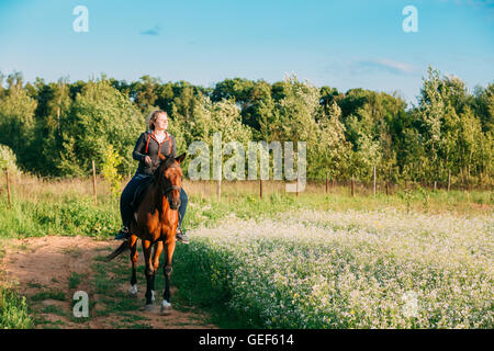 Die junge, lächelnde kaukasischen Frau In Freizeitkleidung braun Reiten entlang der blühenden Wiese Sommerwiese auf sonnigen Land Stockfoto