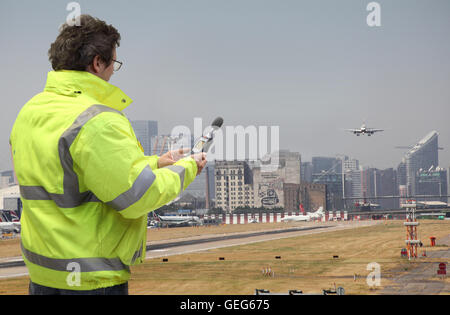 Ein Umwelt-Ingenieur überwacht Lärmpegel am London City Airport Flugzeug landet. Canary Wharf im Hintergrund. Stockfoto