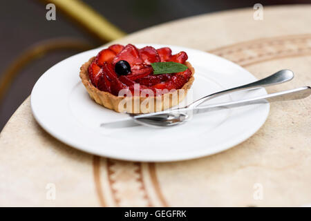 Foto einer Obst-Torte mit Erdbeeren, Minze und schwarzen Johannisbeeren, auf einem weißen Teller mit Messer und Gabel auf einen Tisch im Restaurant. Stockfoto