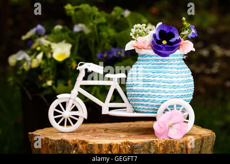 Foto von einem weißen Fahrrad mit lila und rosa Blüten auf Gepäck auf einem Baumstumpf. Stockfoto