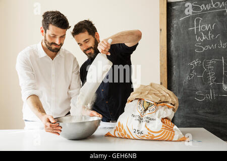 Zwei Bäcker an einem Tisch stehend, Brotteig vorbereiten, aus einer Papiertüte in eine Metall Schüssel Mehl hinzufügen. Stockfoto
