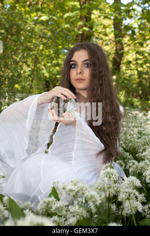 Schöne junge Frau trägt ein weißes Kleid in einem Wald eine Kristallkugel hält Stockfoto