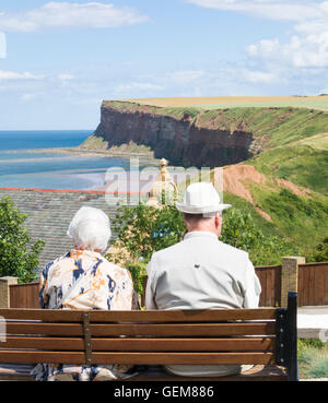 Älteres Ehepaar sitzt auf Sitzplatz mit Blick auf Saltburn Strand und Klippen an einem sonnigen Tag. Saltburn am Meer. Yorkshire, England. UK Stockfoto