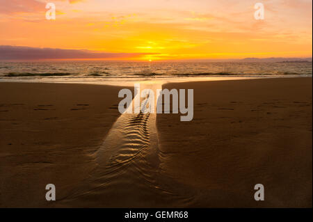 Meer Sonnenuntergang ist ein Sandstrand malerisch mit einem weich fließenden Strom und eine lebendige pulsierende Sonnenuntergang Himmel am Horizont. Stockfoto