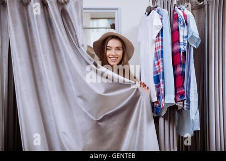 Fröhliche junge Frau mit Hut bedeckt sich stehend in Umkleidekabine