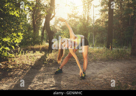 Junge Sportlerin dehnen. Schöne sportliches Mädchen Übungen im Wald bei farbenfrohen Sonnenuntergang im Sommer abends sport. Fitness Stockfoto