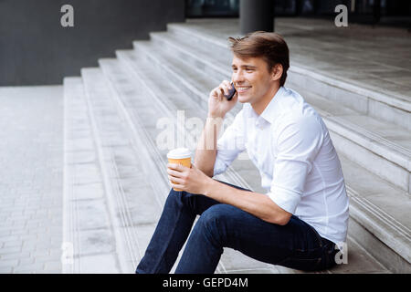 Fröhlicher junger Geschäftsmann mit Tasse Kaffee auf der Treppe sitzen und telefonieren mit Handy Stockfoto