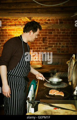 Ein Mann in ein blau-weiß gestreiften Schürze Fisch auf einer heißen Platte auf einem Herd kochen. Stockfoto