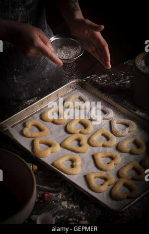 Valentinstag Backen, Frau Beregnung Puderzucker über Herz geformten Kekse auf ein Backblech. Stockfoto