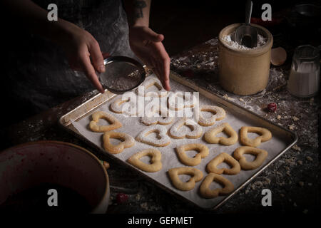 Valentinstag Backen, Frau Beregnung Puderzucker über Herz geformten Kekse auf ein Backblech. Stockfoto