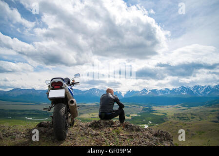 Glatze Fahrer sitzt auf einem Berg neben dem Motorrad und sieht asi Stockfoto