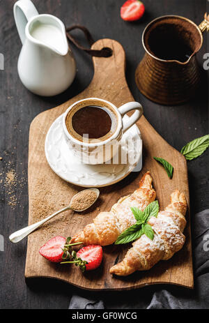 Frühstücks-Set. Frisch gebackene Croissants mit Erdbeere, Minze und Kaffee auf Holzbrett über dunklen Hintergrund Stockfoto