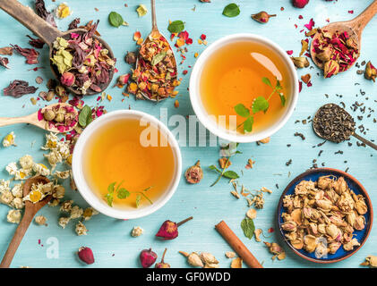Zwei Tassen gesunde Kräuter-Tee mit Minze, Zimt, Trockenblumen Rose und Kamille in Löffel auf blauem Hintergrund Stockfoto
