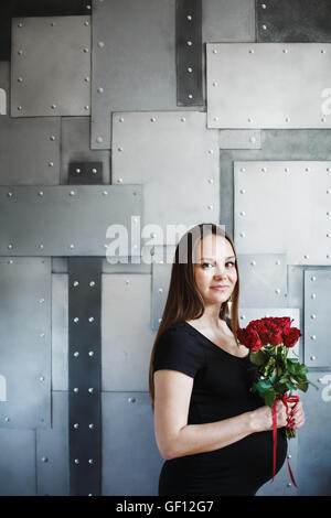 Porträt des eleganten schwangere Frau im schwarzen Kleid mit roten Rosen in der Kamera suchen. Platz für Text. selektiven Fokus. Stockfoto
