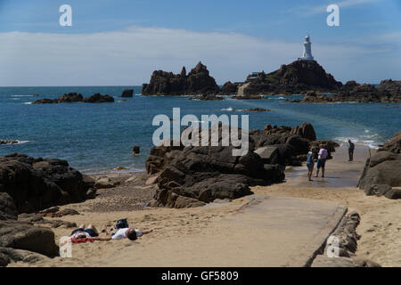 Touristen entspannen Sie sich auf einen Teil des sandigen Strandes mit La Corbiere Leuchtturm im Hintergrund, Jersey, Kanalinseln Stockfoto