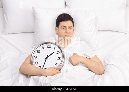 Frustrierte junge Mann im Bett liegend und hält eine Uhr in der Hand isoliert auf weißem Hintergrund Stockfoto