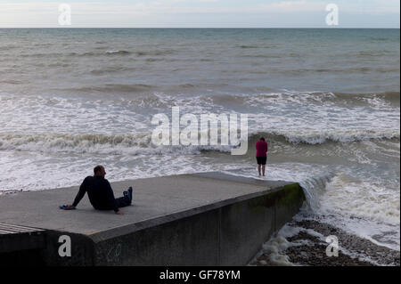 Mann sitzt auf einem Wellenbrecher, die gerade seine Frau Paddeln im Meer, Normandie, Frankreich Stockfoto