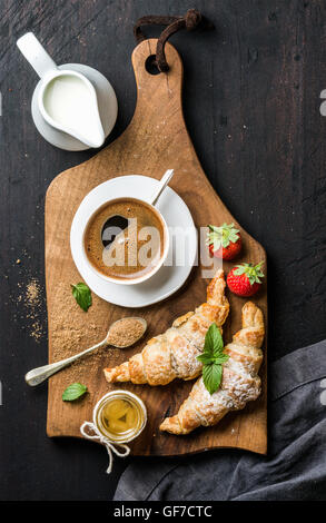Frühstücks-Set. Zwei frisch gebackene Croissants mit Erdbeeren, Honig, schwarzer Kaffee, Krug und Löffel auf rustikalem Holzbrett auf dunklem Hintergrund Stockfoto