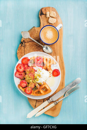 Frühstücks-Set. Warme hausgemachte belgischen Waffeln mit Sahne, Erdbeeren, Ahornsirup und zerkleinerten Pistazien, Tasse Espresso, brauner Zucker auf Oliven rustikalen Holzbrett über blau bemalten Hintergrund Stockfoto