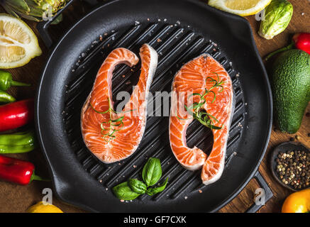 Abendessen kochen Zutaten. Zwei Stücke von rohen rohen Lachs mit Gemüse, Kräutern, Zitrone, Avocado, Artischocken, Eisen Gewürze Grillen Pfanne über hölzerne Hintergrund Stockfoto