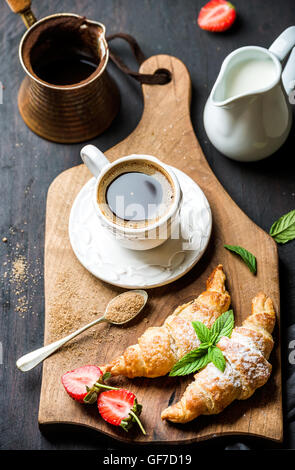 Frühstücks-Set. Frisch gebackene Croissants mit Erdbeere, Minze und Kaffee auf Holzbrett über dunklen Hintergrund Stockfoto