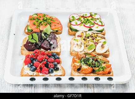 Süßes und herzhaftes Frühstück Toast Vielfalt. Sandwiches mit Obst, Gemüse, Eiern, geräucherter Lachs auf Backblech in hellem Holz Hintergrund weiß Stockfoto