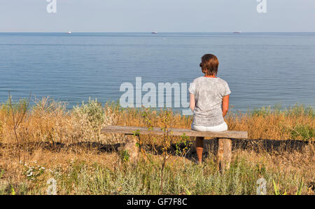 weiße mittlere gealterte Frau mit kurzen Haaren in T-shirt, Shorts und Sonnenbrille sitzt auf alten Holzbank mit schwarzen Meer Meeresblick Stockfoto