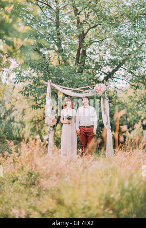 Der Bräutigam Hochzeitsanzug und Braut trägt Spitze Kleid stehen, Hand in Hand, Bogen auf Hintergrund Stockfoto