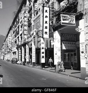 Historische, ruhige Seitenstraße der 1950er Jahre in Hongkong, die ein großes Mehrzweckbetongebäude mit überdachtem Bürgersteig oder Gehweg neben den Geschäften oder Einheiten im Erdgeschoss zeigt, mit zahlreichen Schildern mit chinesischen Schriftzeichen, die die tragenden Betonpfosten und die Außenstruktur des Gebäudes bedecken. In der nahe leeren Straße werden ein Solidaritäts-Motorwagen und -Motorrad geparkt, wobei die Hintereingänge und Warenlieferbereiche für die Geschäfte vorhanden sind. Stockfoto