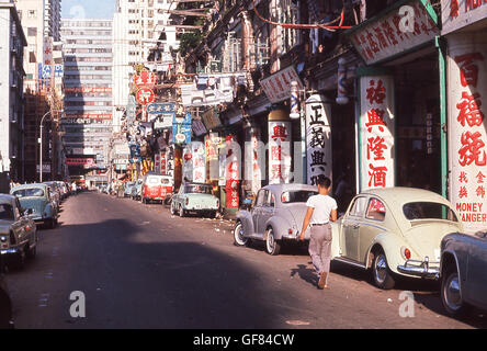1960er, historisch, tagsüber und Blick auf eine Seitenstraße in der Altstadt von Hongkong, mit chinesischen Schriftzeichen, die die Betonpfosten der Straßengebäude bedeckten. Autos der Ära stehen daneben. Stockfoto