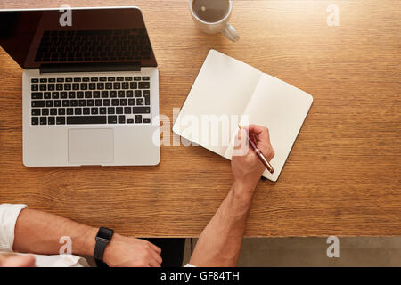 Nahaufnahme des Menschen, die Schreiben von Notizen in persönliches Notizbuch mit ein Laptop und eine Tasse Kaffee am Tisch. Stockfoto