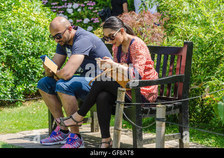 Ein paar sitzen zusammen auf einer Bank im Holland Park, London und nutzen ihre Smartphones. Stockfoto