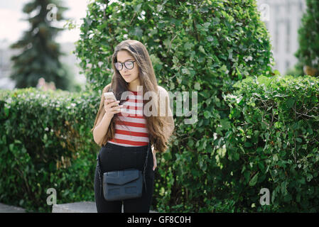 Junge asiatische Frau mit Brille mit Smartphone und Sms senden. Junge Frau auf Stadt-Hintergrund mit grünen Blättern. Stockfoto