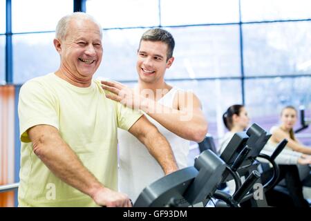 EIGENTUM FREIGEGEBEN. -MODELL VERÖFFENTLICHT. Junger Mann unterstützt ältere Menschen während dem Training auf dem Heimtrainer im Fitness-Studio. Stockfoto