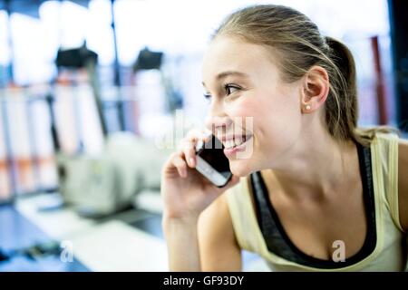 EIGENTUM FREIGEGEBEN. -MODELL VERÖFFENTLICHT. Junge Frau lächelt und telefonieren mit Handy im Fitness-Studio. Stockfoto