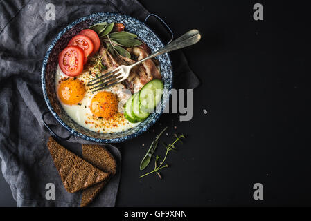 Frühstücks-Set. Pfanne mit Spiegelei mit Speck, frische Tomaten, Gurken, Salbei und Brot auf dunklen Portion Brett auf schwarzem Hintergrund Stockfoto