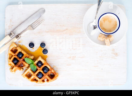 Espresso-Kaffee-Tasse, weiche belgische Waffeln mit frischen Heidelbeeren und Marple Sirup auf weiß lackiertem Holzbrett auf hellblauem Hintergrund Stockfoto