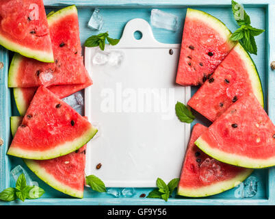 Wassermelone Scheiben mit Eiswürfeln und Minze auf blauen hölzernen Hintergrund, weiße Keramik Board im Zentrum. Ansicht von oben, Textfreiraum Stockfoto