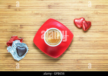 Herzförmige Bonbons eingewickelt in einer leuchtend roten Folie auf Holz Textur mit Tasse Kaffee in der Nähe liegen. Hintergrund für romantische Themen. Stockfoto