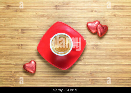 Herzförmige Bonbons eingewickelt in einer leuchtend roten Folie auf Holz Textur mit Tasse Kaffee in der Nähe liegen. Hintergrund für romantische Themen. Stockfoto