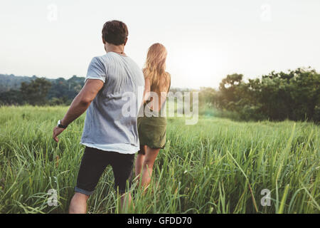 Rückansicht eines jungen Paares auf einer Wiese spazieren. Junger Mann und Frau zusammen in ländlichen Landschaft spazieren. Stockfoto