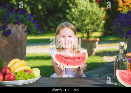 Glückliches Mädchen mit großen roten Scheibe Wassermelone auf rustikale Tabellen im Sommergarten sitzen. Stockfoto