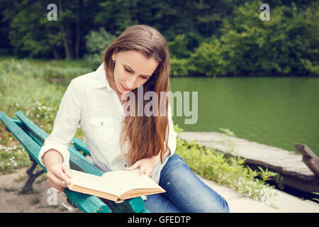 Junge Frau im Park in der Nähe von Teich und Lesung Buch. Ziemlich kaukasische Mädchen mit Buch entspannen auf der Bank Stockfoto