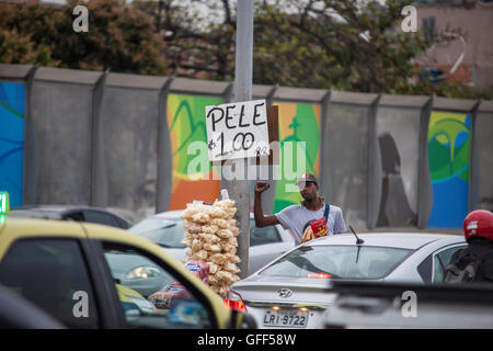 Bewohner von Complexo da Mare, einer massiven Netzwerk der Favelas, die neben der Linha Vermelha (rote Linie), die Autobahn vom Flughafen in die Innenstadt, Rio De Janeiro sitzt arbeiten als Straßenverkäufer während der Stoßzeiten an der Expreessway - seit 2010 ist die Gemeinschaft eingezäunten Weg von der Autobahn durch riesige Plexiglas Platten - Behörden behaupten sie bieten eine akustische Barriere , einheimischen beschreiben es als eine "Mauer der Schande", anders Arme Menschen zu verstecken. Kurz vor den Olympischen Spielen begann die Stadt Verputzen die 3 Meter hohen, 7km langen Tafeln mit bunten Olympia-Plakate s Stockfoto