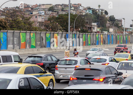 Bewohner von Complexo da Mare, einer massiven Netzwerk der Favelas, die neben der Linha Vermelha (rote Linie), die Autobahn vom Flughafen in die Innenstadt, Rio De Janeiro sitzt arbeiten als Straßenverkäufer während der Stoßzeiten an der Expreessway - seit 2010 ist die Gemeinschaft eingezäunten Weg von der Autobahn durch riesige Plexiglas Platten - Behörden behaupten sie bieten eine akustische Barriere , einheimischen beschreiben es als eine "Mauer der Schande", anders Arme Menschen zu verstecken. Stockfoto