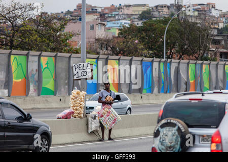 Bewohner von Complexo da Mare, einer massiven Netzwerk der Favelas, die neben der Linha Vermelha (rote Linie), die Autobahn vom Flughafen in die Innenstadt, Rio De Janeiro sitzt arbeiten als Straßenverkäufer während der Stoßzeiten an der Expreessway - seit 2010 ist die Gemeinschaft eingezäunten Weg von der Autobahn durch riesige Plexiglas Platten - Behörden behaupten sie bieten eine akustische Barriere , einheimischen beschreiben es als eine "Mauer der Schande", anders Arme Menschen zu verstecken. Stockfoto