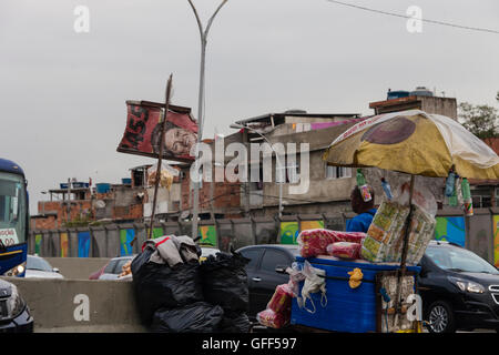 Bewohner von Complexo da Mare, einer massiven Netzwerk der Favelas, die neben der Linha Vermelha (rote Linie), die Autobahn vom Flughafen in die Innenstadt, Rio De Janeiro sitzt arbeiten als Straßenverkäufer während der Stoßzeiten an der Expreessway - seit 2010 ist die Gemeinschaft eingezäunten Weg von der Autobahn durch riesige Plexiglas Platten - Behörden behaupten sie bieten eine akustische Barriere , einheimischen beschreiben es als eine "Mauer der Schande", anders Arme Menschen zu verstecken. Stockfoto