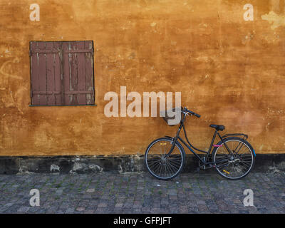 Ein einsames Fahrrad die verwitterten orange Wand eines Hauses mit einem ebenso verwitterten geschlossenem Verschluss gelehnt. Stockfoto