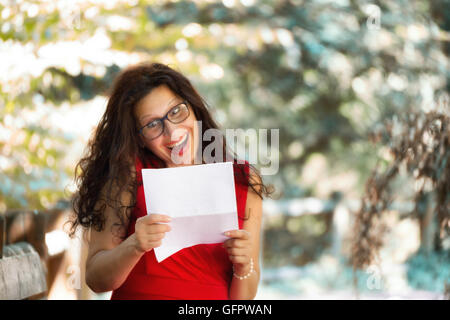 wunderschöne Frau mittleren Alters in einem roten Kleid und langen braunen welliges Haar ist eine Zeitung lesen und Lachen mit einer Grimasse karikierte Begeisterung beim Tragen einer Brille nerdy in einem Garten Stockfoto