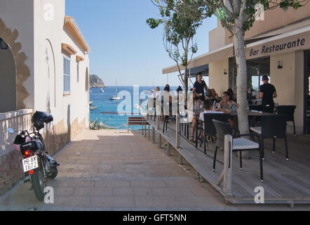 Restaurant mit Blick auf das Meer mit Menschen und geparkten Motorrad in Sant Elm an einem sonnigen Sommertag im Juli. Stockfoto